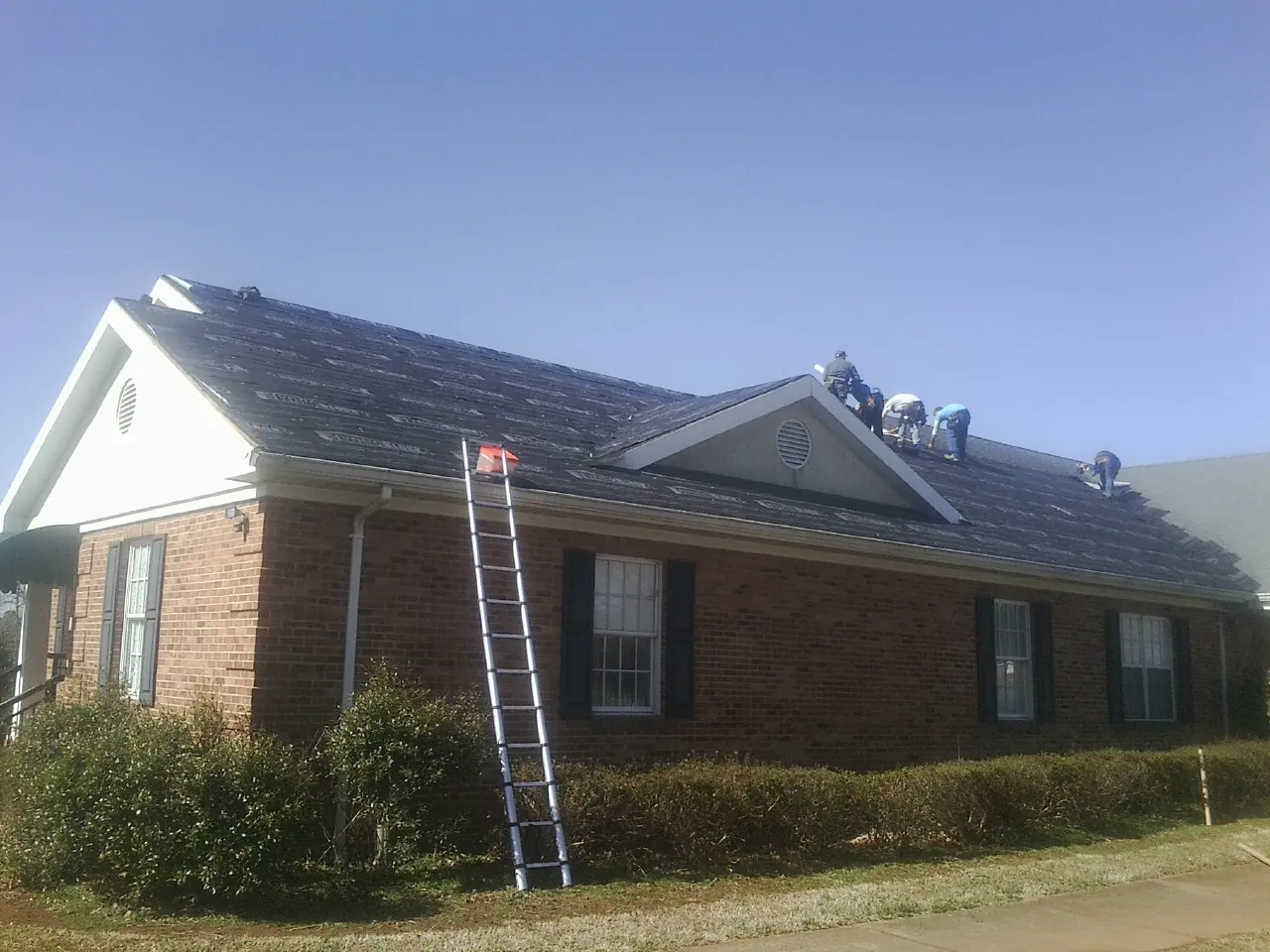Crew tearing shingles off a brick ranch roof with a ladder set — Emergency Roof Repair work by our crew in Lane