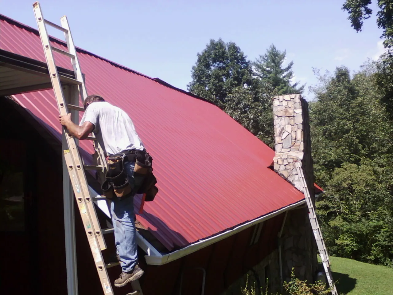 Roofer on a ladder beside a red metal roof and stone chimney — Soffit & Fascia Repair by Roofing Brothers in Lane